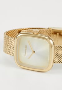 Montre avec un bracelet en maille dorée, un boîtier carré, un cadran métallique clair, des index horaires dorés, et une couronne sur le côté. Marque visible sur le cadran.