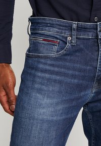 Tmavě modré džínové kalhoty s vybledlou texturou, s klasickým designem pěti kapes a malým červeným akcentem na zadní kapse.