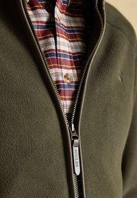 Olivovo zelená fleecová bunda čiastočne zapnutá nad červenou, bielou, modrou a žltou károvanou košeľou s gombíkmi.