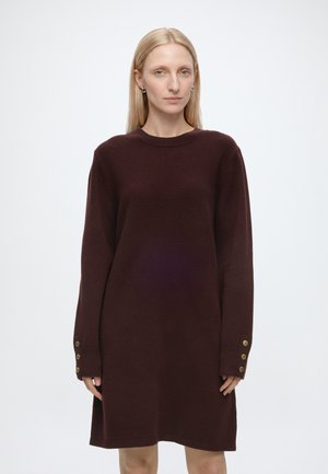 Femme blonde portant une robe-pull marron foncé à manches longues avec des boutons dorés aux poignets, debout devant un fond clair uni.