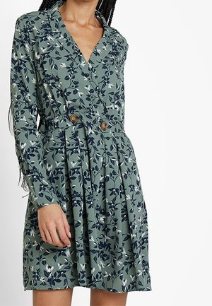Zielona sukienka w kwiaty z kołnierzykiem, długimi rękawami i dopasowaną talią. Ma zapięcie na guziki oraz plisowaną spódnicę z granatowymi akcentami.