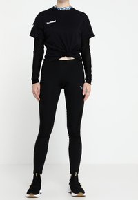 Camiseta atlética negra con nudo en el frente, superpuesta a una blusa de manga larga con estampado. Leggings negros y zapatillas deportivas negras con acentos dorados.