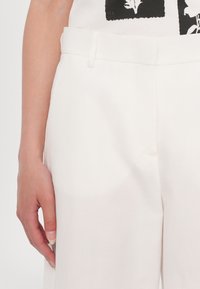 Main gauche reposant à côté d'un pantalon blanc taille haute avec passants pour ceinture, associé à un top blanc et noir imprimé.