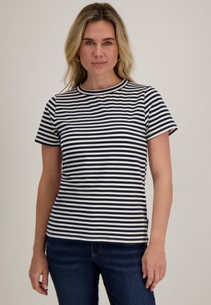 Vrouw met blond haar die een marineblauw-wit gestreept shirt met korte mouwen en donkerblauwe spijkerbroek draagt, staand tegen een effen achtergrond.