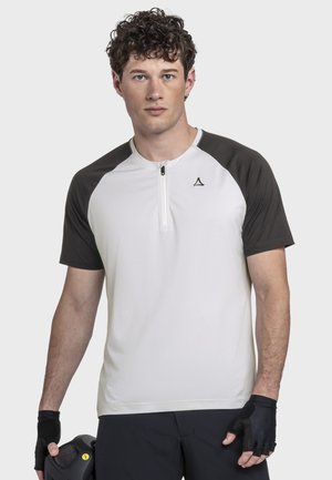 Junger Mann mit lockigem Haar, der ein zweifarbiges Radtrikot und schwarze fingerlose Handschuhe trägt und einen schwarzen Fahrradhelm hält.