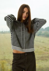 Szary sweter w paski z kapturem, o luźnym kroju i prążkowanym wykończeniu na dole, zestawiony z ciemnobrązowymi spodniami. Materiał wydaje się miękki i przytulny.