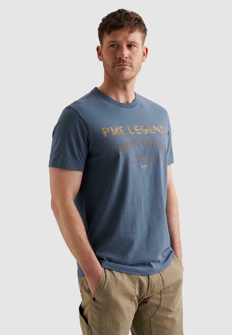 Άνδρας με μπλε μπλούζα PME Legend Cargo Goods και μπεζ παντελόνι cargo, στέκεται με τα χέρια στις τσέπες, κοιτώντας προς τα δεξιά.