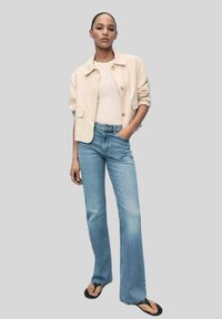 Giacca cropped beige chiaro sopra una maglietta bianca, abbinata a jeans a zampa d'elefante blu chiaro e sandali neri con dettagli decorativi.