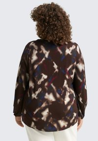 Vrouw met krullend haar in een lange mouw, abstract patroon in bruin, wit, blauw en rood shirt, gezien van achteren.