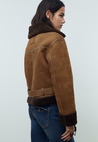 Veste en daim marron avec un col et des poignets en shearling, dotée d'une ceinture à la taille et d'un design court. Portée avec un jean bleu.