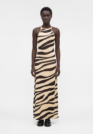 Γυναίκα στέκεται φορώντας ένα μακρύ, μπεζ αμάνικο φόρεμα με μαύρες ρίγες ζέβρας και μαύρα παπούτσια μπροστά σε απλό λευκό φόντο.