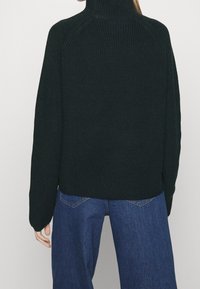 Donkergroene geribbelde coltrui met lange mouwen; heeft een losse pasvorm en zijsplitten, gecombineerd met blauwe jeans met wijd uitlopende pijpen.