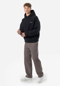 Un sweat à capuche noir en polaire avec une poche kangourou et un logo violet, associé à un pantalon gris clair et des baskets blanches, sur un fond blanc uni.