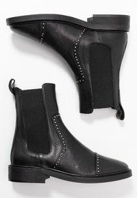 Botas negras de cuero hasta el tobillo con lados elásticos, acentos de tachuelas a lo largo de las costuras y suela plana. Presenta una punta redondeada y una pestaña de agarre.