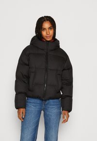 Schwarze Steppjacke mit hohem Kragen und Reißverschluss, mit gestepptem Design und Seitentaschen. Getragen über einem roten Oberteil und blauen Jeans.
