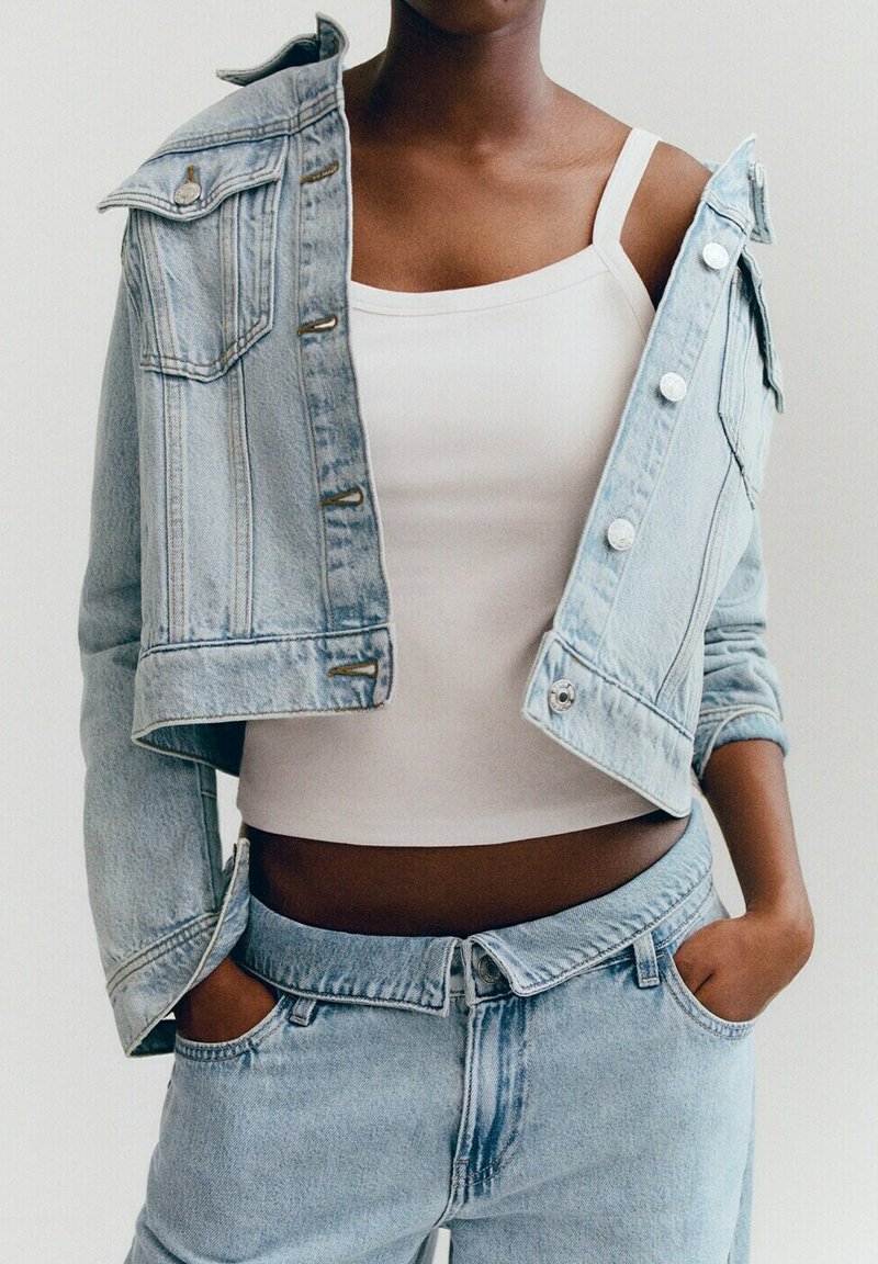 Person trägt ein weißes Tanktop und eine hellblaue Jeansjacke, die über eine Schulter gezogen ist, kombiniert mit passenden hellblauen Jeans.