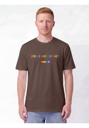 Mann mit roten Haaren, der ein braunes T-Shirt mit dem Text "Spread Love Not Hate" in Regenbogenfarben und sechs kleinen bunten Herzen unter dem Text trägt.