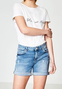 White short-sleeve t-shirt with black script text, paired with light blue denim shorts featuring rolled cuffs and distressed details.