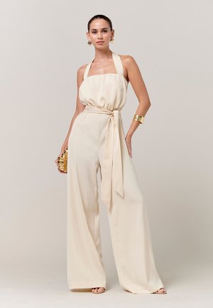 Kobieta ubrana w kremowy kombinezon typu halter z szerokimi nogawkami, trzymająca złotą kopertówkę, ozdobiona złotymi kolczykami i bransoletkami.