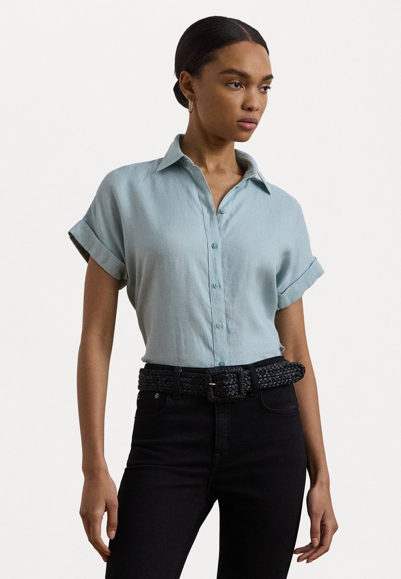 Ανοιχτό μπλε λινό πουκάμισο με γιακά και γυρισμένα μανίκια, σε συνδυασμό με ψηλόμεσα μαύρα τζιν και μία υφή μαύρη πλεχτή ζώνη.