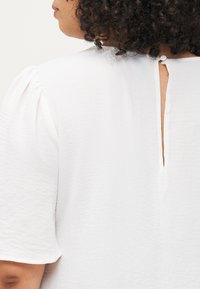 White short-sleeve blouse featuring a textured fabric, round neckline, and a back closure with a small button and slit detail.