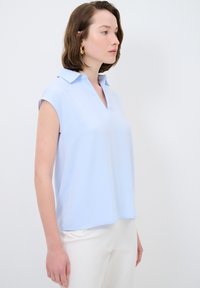 Blusa blu chiaro a maniche corte con collo e vestibilità morbida. Tessuto liscio e leggero con una leggera lucentezza.