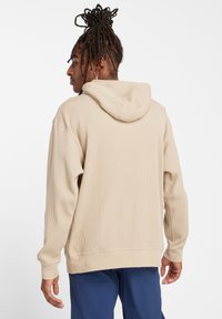 Beige ribbed hoodie with a lined hood, kangaroo pocket, and relaxed fit, featuring textured fabric and ribbed cuffs and hem.