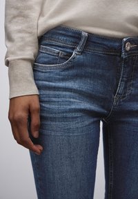 Jeans de mezclilla azul con un corte ajustado, que cuentan con un diseño tradicional de cinco bolsillos, costuras visibles y un cierre de botón metálico.