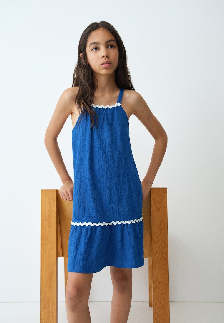 Jeune fille portant une robe bleue sans manches avec bordure blanche, debout les mains sur les hanches devant une chaise en bois sur fond blanc uni.