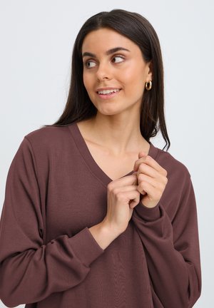 Ung kvinde med langt mørkt hår iført en brun V-hals sweater og gyldne hoop øreringe, der ser til siden med hænderne foldet tæt ved brystet.