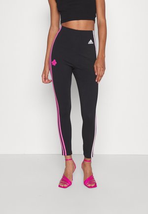 Mujer con leggings negros de Adidas con rayas laterales rosas y detalle floral, combinados con sandalias de tacón alto rosas y un top corto negro.