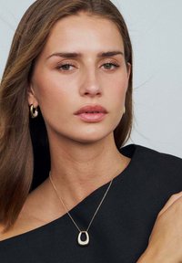 Guld halskæde med en dråbeformet vedhæng, båret med en sort off-shoulder top. Håret er langt og brunt med subtile bølger. Enkle hoops øreringe.