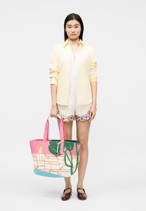 Kobieta w jasnożółtej koszuli na guziki, białych szortach z haftem kwiatowym, brązowych butach typu Mary Jane, trzymająca dużą, kolorową torbę.