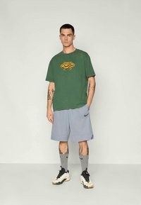 Grønn t-skjorte med korte ermer og en gul logo på fronten, kombinert med lysegrå treningshorts og sorte joggesko med hvite detaljer.