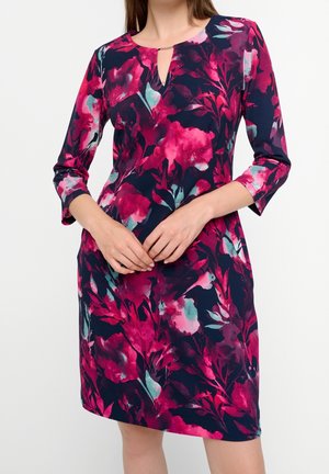 Femme portant une robe mi-longue avec un motif floral rose, rouge et bleu sarcelle sur fond bleu foncé et manches trois-quarts.