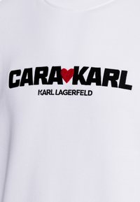 Sudadera blanca con texto bordado en negro y rojo: "CARA♥KARL" con "KARL LAGERFELD" en letras más pequeñas debajo. Textura suave.