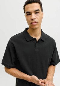 Homme aux cheveux courts et bouclés portant un polo noir côtelé, regardant la caméra avec les mains jointes devant lui.