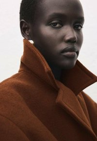 Bruine wollen jas met een hoge kraag en een uitgesproken revers, met een zachte textuur. Model draagt gouden oorringen en neutrale make-up.