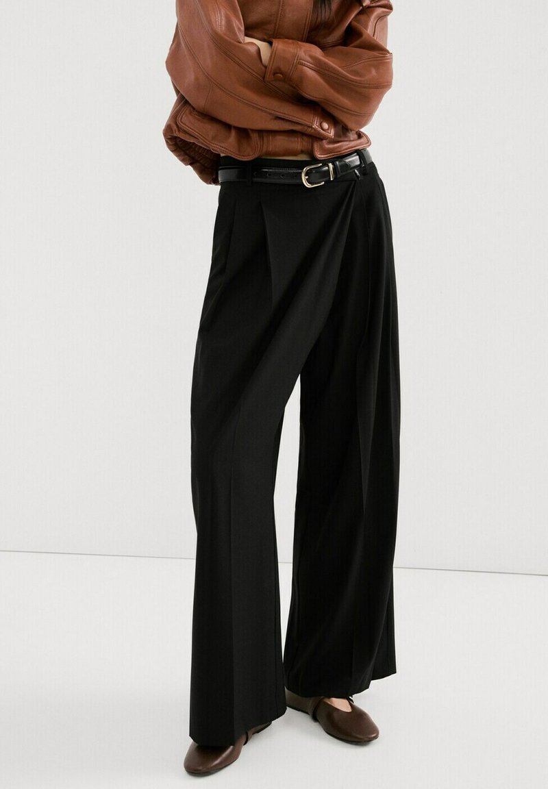 Person trägt schwarze Hosen mit weitem Bein, braunen Ledergürtel, braune Schuhe und braune Jacke, die Arme verschränkt.