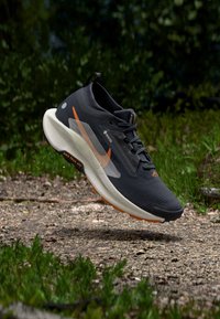 Schwarzer Sportschuh mit strukturiertem Obermaterial, Mesh-Einsatz, orangefarbenen Akzenten und weißer Zwischensohle, schwebend über einem Kiesweg mit Grünbewuchs.