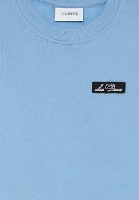 Light blue sweatshirt with a round neckline, ribbed cuffs, and a sewn black label featuring the text "Les Deux." Smooth cotton fabric.