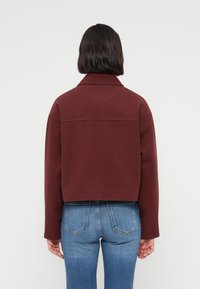 Κοντό μπορντό σακάκι με δομημένο γιακά, μακριά μανίκια και λεία υφή. Φορεμένο πάνω από μπλε τζιν με ορατά πίσω τσέπες.