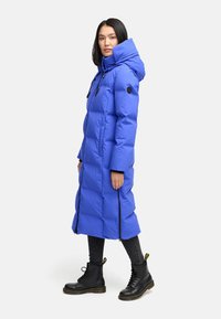 Lange blauwe pufferjas met een capuchon, voorzien van een ritssluiting, zijzakken en contrasterende zwarte accenten. Draagt het met zwarte combatlaarzen.
