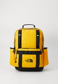 Gul nylon rygsæk med sorte detaljer, flere rum, polstrede stropper og et top håndtag. Har logo-tryk på forsiden.