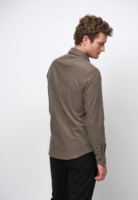 Koszula z długim rękawem w oliwkowym kolorze, wykonana z miękkiego materiału. Posiada prosty kołnierzyk, mankiety zapinane na guziki oraz zaokrąglony dół. Noszona z czarnymi spodniami.