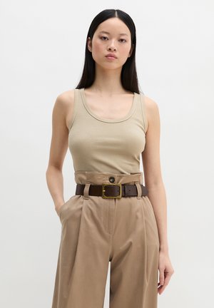 Frau mit langem schwarzen Haar, die ein beiges geripptes Tanktop und hoch taillierte khakifarbene Hose mit einem dunkelbraunen Gürtel trägt, steht mit einer Hand in der Tasche.