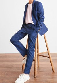 Marineblauw pak met een getailleerde pasvorm, gecombineerd met een lichtroze t-shirt en witte sneakers. Houten kruk en houten vloer op de achtergrond.