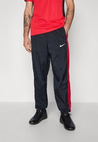 Pantalons de sport Nike noirs avec des bandes latérales rouges, fabriqués en tissu léger et associés à des baskets noires. Texture lisse et coupe décontractée.