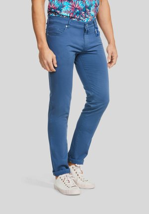 Man draagt slim-fit blauwe broek gecombineerd met witte sneakers en een tropisch overhemd, staand tegen een effen achtergrond.
