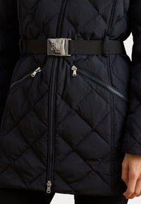 Must mustrõngaga must tikkpuffer jope, millel on kuldne logo pandla, kaks tõmblukk-taskut ja tekstureeritud teemantmuster.